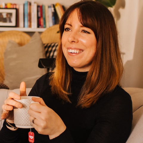 A smiling white woman with long dark hair sitting on the sofa in a cosy room with a nice cup of tea.
