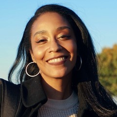 An outdoor portrait of a smiling dark-skinned woman smiling with shoulder-length black hair.