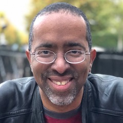 A smiling black man with glasses and close-cropped hair and beard wearing a leather jacket outdoors.