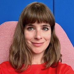 A smiling white woman with shoulder-length brown hair wearing a bright red top in a pink chair in front of a bright blue wall.