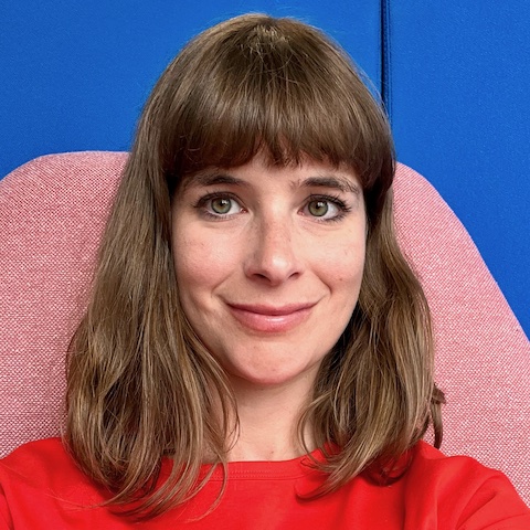 A smiling white woman with shoulder-length brown hair wearing a bright red top in a pink chair in front of a bright blue wall.