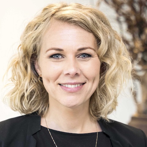 A smiling woman with wavy blonde hair, pale skin and light blue eyes wearing a dark outfit in front of a light background.