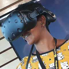 A brown-skinned woman with short hair and a colourful yellow top wearing a virtual reality headset looking to one side.